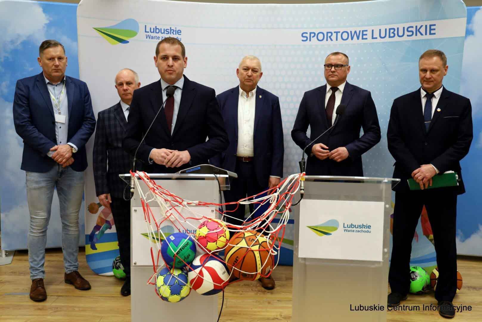 Na pierwszym planie mężczyzna stoi przy mównicy W tle sotją osoby na tle ścianki  Miliony na turystykę boiska i pumptracki Zyskały małe gminy WIDEO