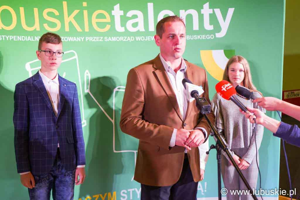 Mężczyzna i dwie młode osoby na tle zielonej planszy