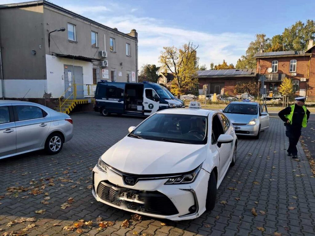 Na zdjęciu dwie taksówki. Obok jednej z nich stoi policjant. W tle widać auta mundurowych.