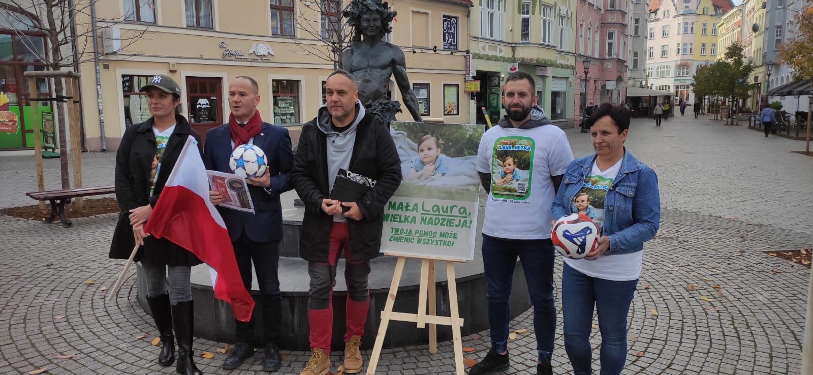 Uczestnicy konferencji zapowiadającej memoriał cieja stoją na tle pomniku Bachusa na zielonogórskim deptaku  Biało czerwoni zagrają dla Laury Wolniewicz