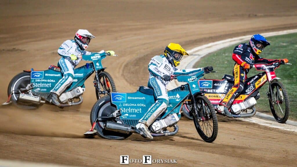 Trzy osoby ścigające się na motocyklach żużlowych