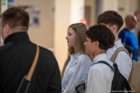 Group of young adults in a hallway, a woman with long blond hair in a white blouse stands in profile.