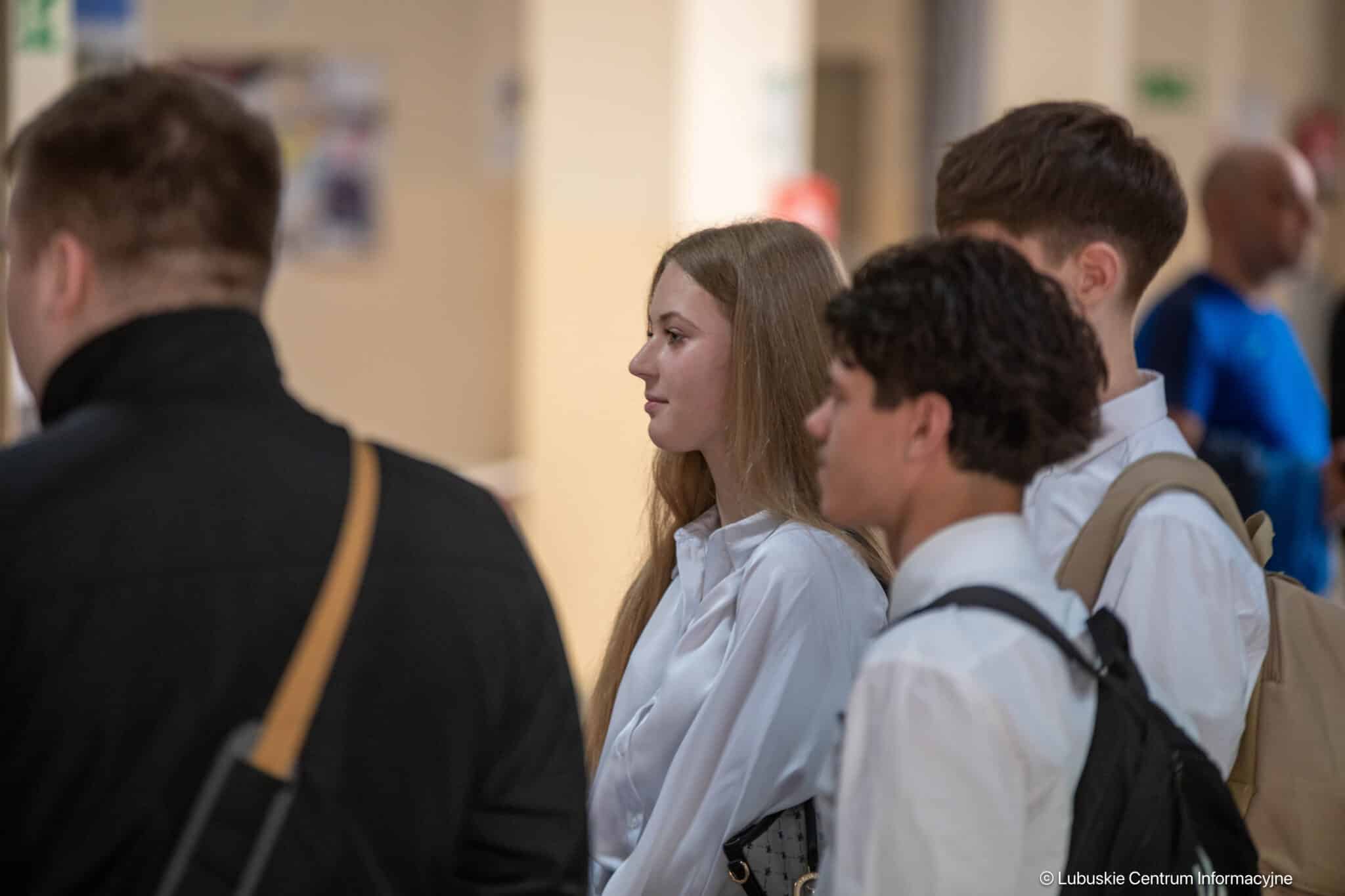 Group of young adults in a hallway, a woman with long blond hair in a white blouse stands in profile.