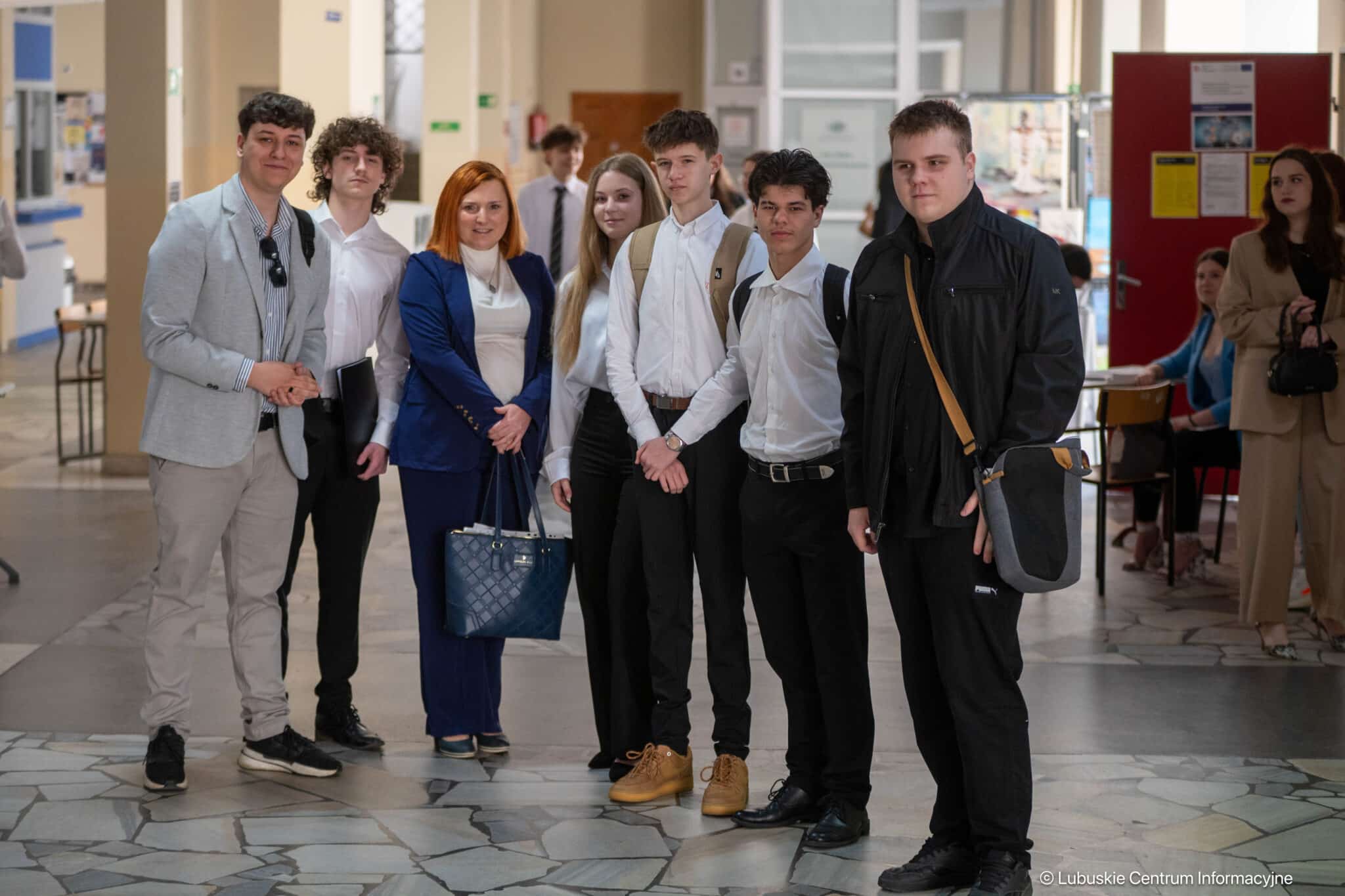 Group of students and a woman in a blue suit posing for a photo in a school hallway; everyone facing camera with backpacks and bags nearby.