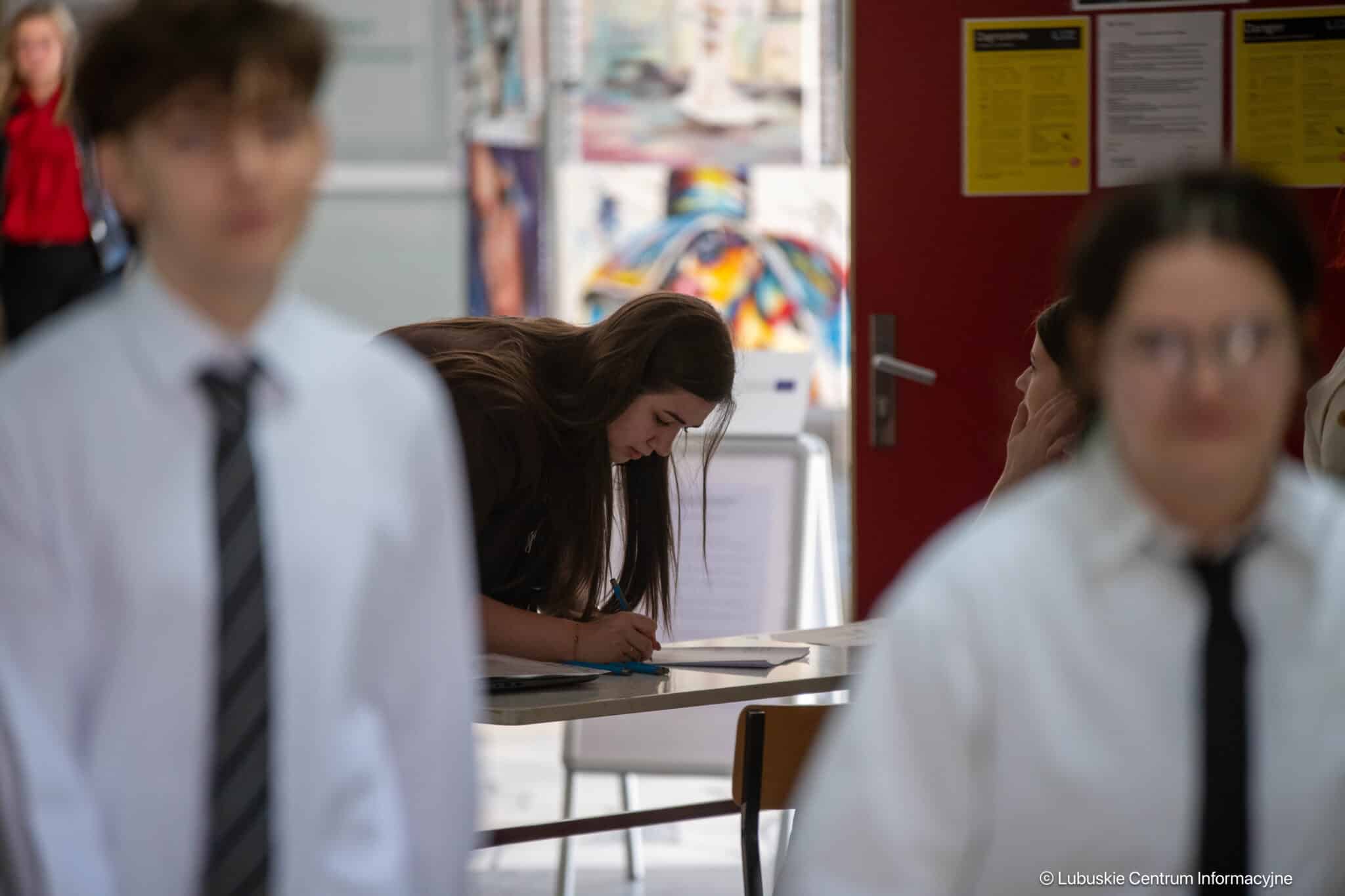 Student leaning over a desk, writing in a school hallway with blurred classmates in the foreground.