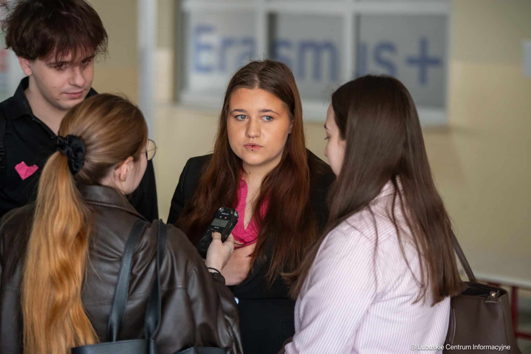 Group of four young adults in a hallway; a woman in the center is being interviewed while others listen, one holds a voice recorder close to her.