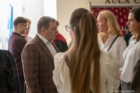 Group of adults chatting in a hallway during an event, a man in a brown plaid blazer converses with women in white blouses to the right.