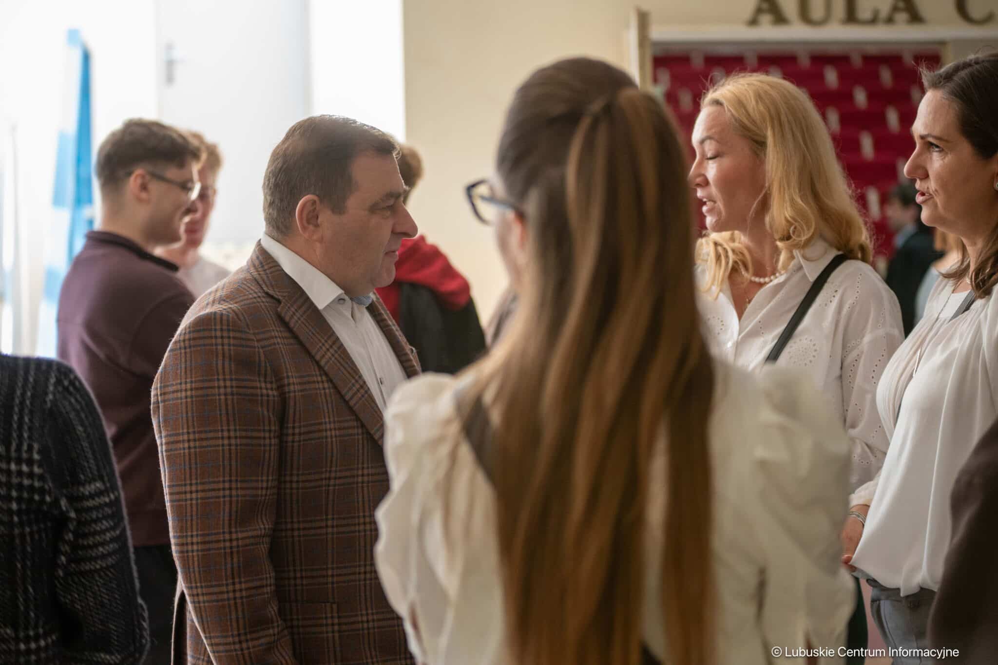 Group of adults chatting in a hallway during an event, a man in a brown plaid blazer converses with women in white blouses to the right.