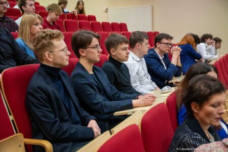 Several young adults seated in a university lecture hall with red upholstered chairs, listening attentively.