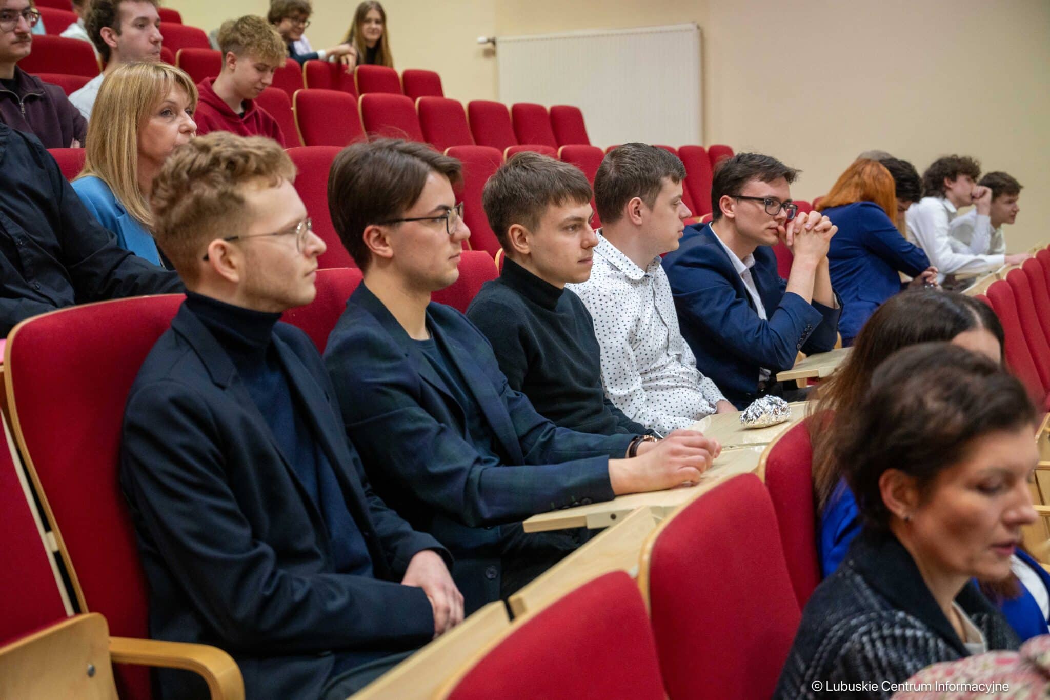 Several young adults seated in a university lecture hall with red upholstered chairs, listening attentively.
