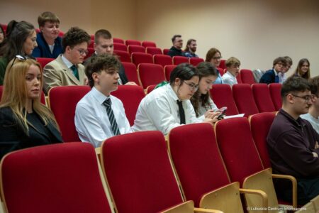 Students sit in a lecture hall with red upholstered chairs, some taking notes while others look at phones or chat nearby.