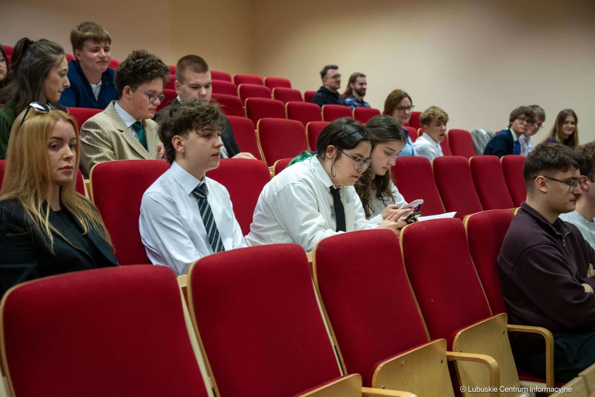 Students sit in a lecture hall with red upholstered chairs, some taking notes while others look at phones or chat nearby.