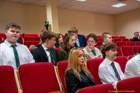 Group of students in formal attire seated in a red-upholstered lecture hall, some chatting.