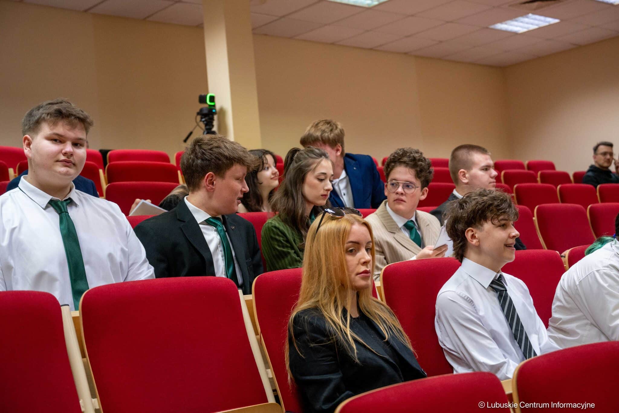 Group of students in formal attire seated in a red-upholstered lecture hall, some chatting.
