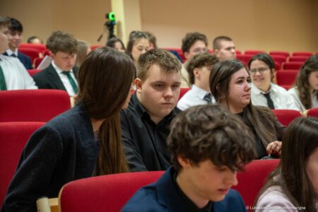 Group of students seated in a lecture hall with red chairs, some conversing in the foreground.