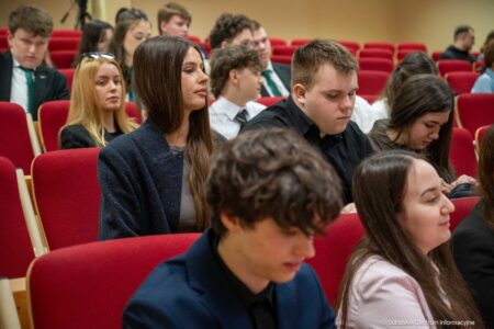 Group of young adults seated in a lecture hall with red chairs, paying attention to an event.