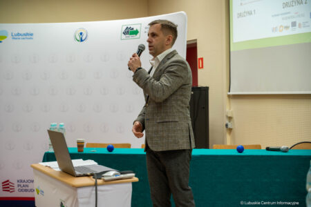 Man in a checked blazer speaks into a handheld microphone at a conference, standing beside a laptop on a table with water bottles and notes behind.