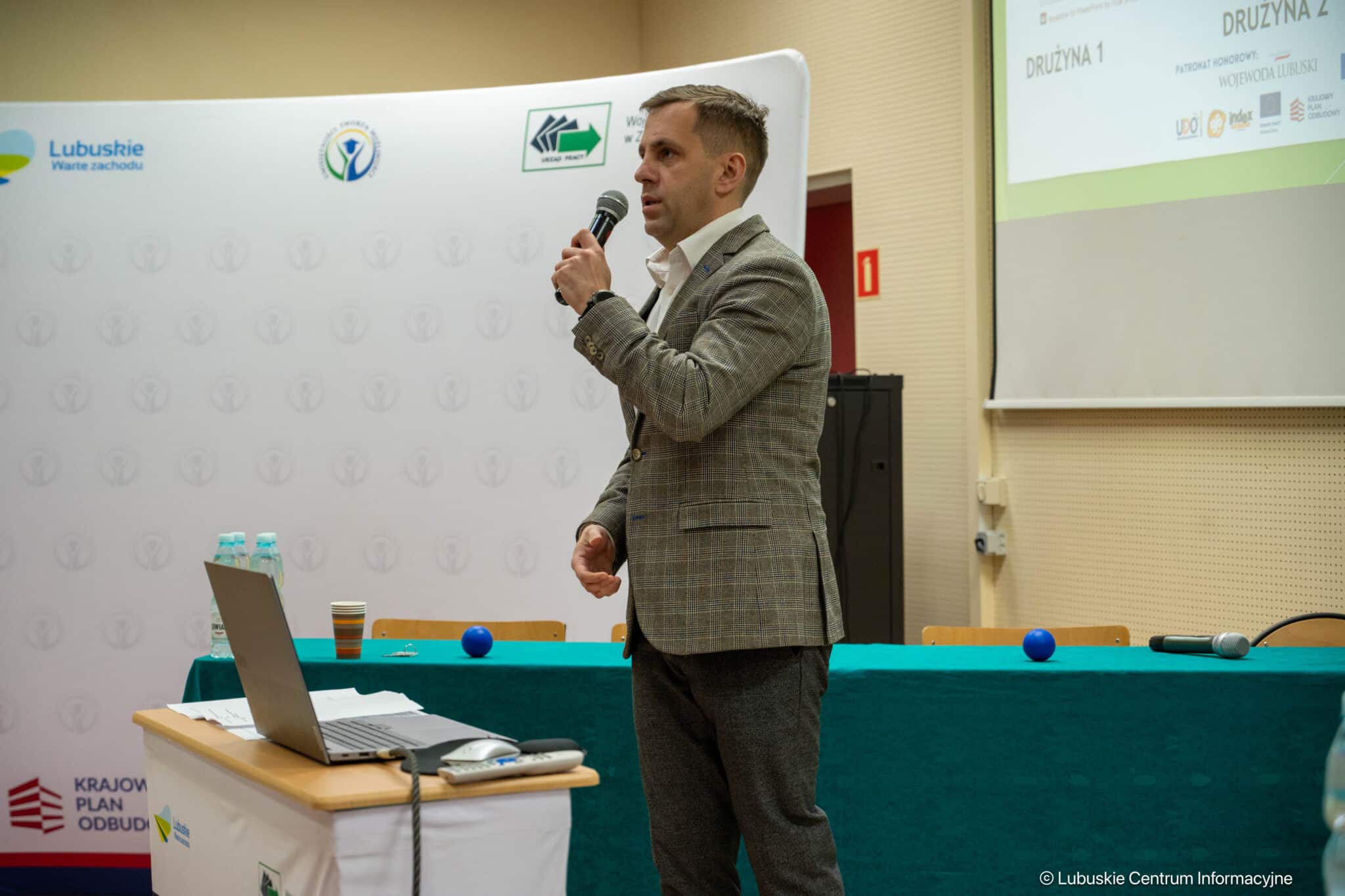 Man in a checked blazer speaks into a handheld microphone at a conference, standing beside a laptop on a table with water bottles and notes behind.