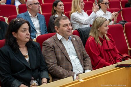 Audience members seated in red theater chairs at a formal event, attention focused forward; man in a brown plaid blazer center with two women nearby.