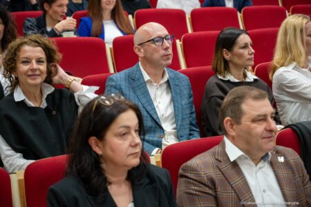 Audience members seated in red upholstered chairs at a conference, attentively listening to a presentation on stage.