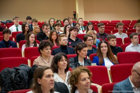 Audience seated in a red-carpeted lecture hall, watching a presentation attentively.