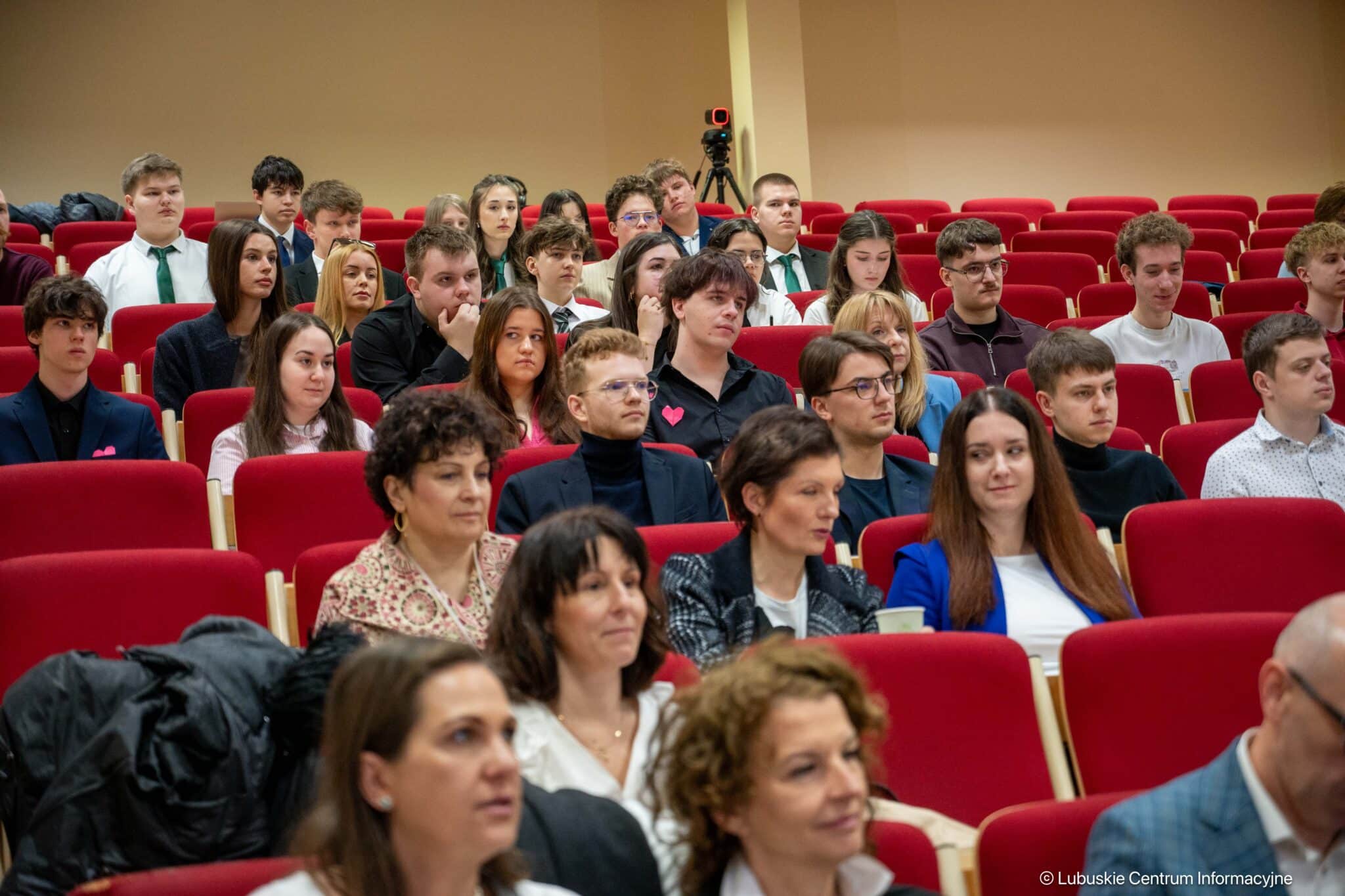 Audience seated in a red-carpeted lecture hall, watching a presentation attentively.