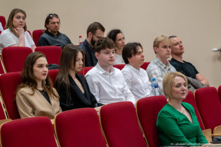Audience of young adults seated in red auditorium chairs listening to a presentation in a conference setting.