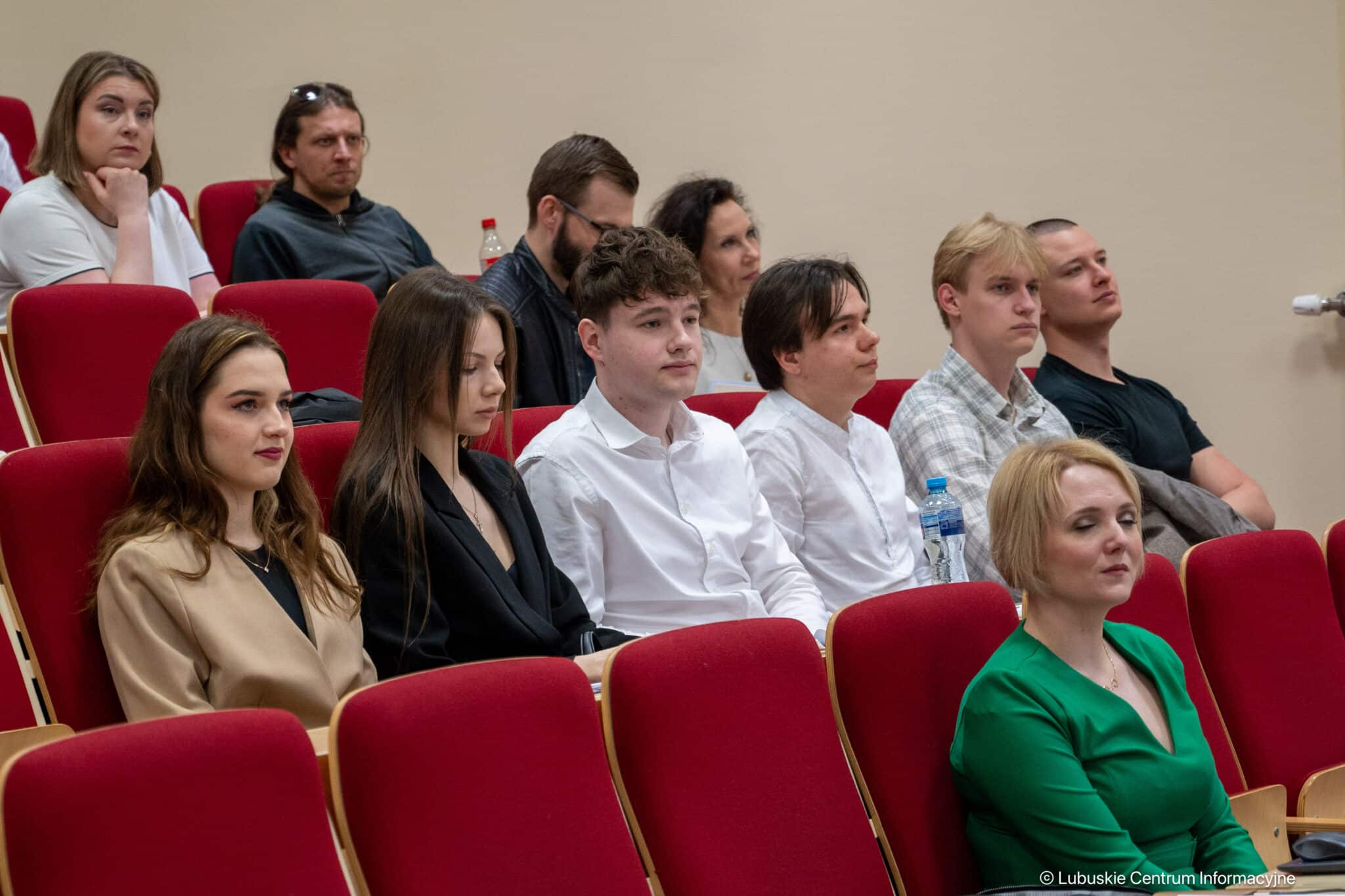 Audience of young adults seated in red auditorium chairs listening to a presentation in a conference setting.