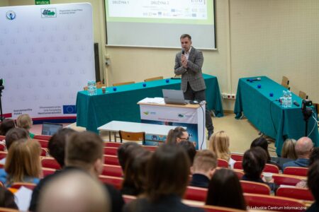 Man in a gray blazer speaks into a microphone at a conference podium in a lecture hall, with a projector screen and banners in the background.