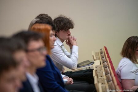 Young man with curly hair in a white shirt sits with his hands folded near his mouth, listening in a classroom.