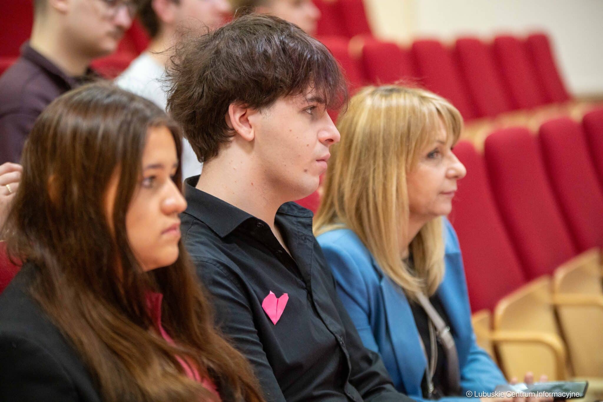 Three people seated in an auditorium, listening to a talk, with red seats in the background.