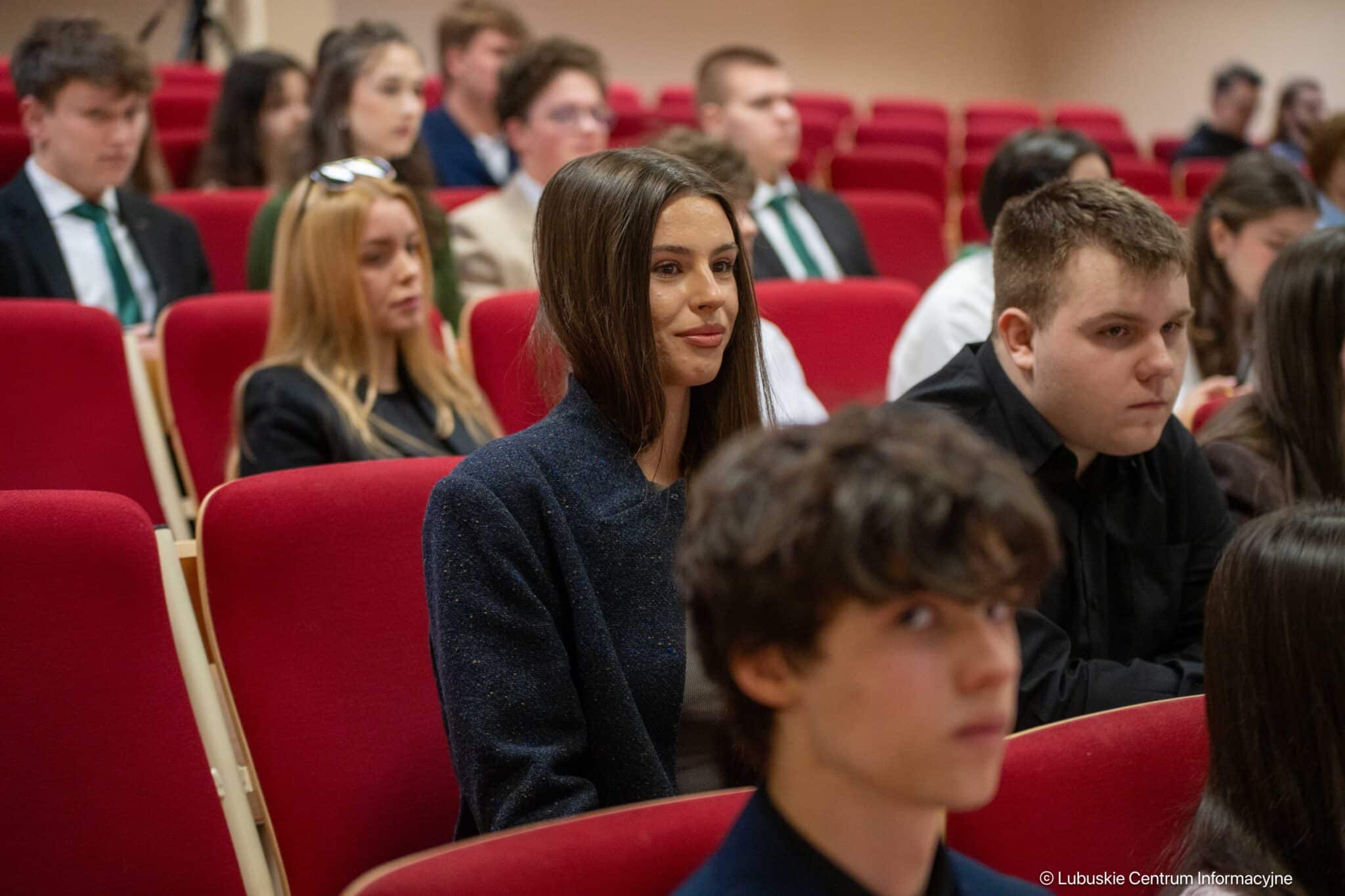 Woman with shoulder-length dark hair sits in a red lecture hall seat, attentive during a talk; other attendees blurred in the background.