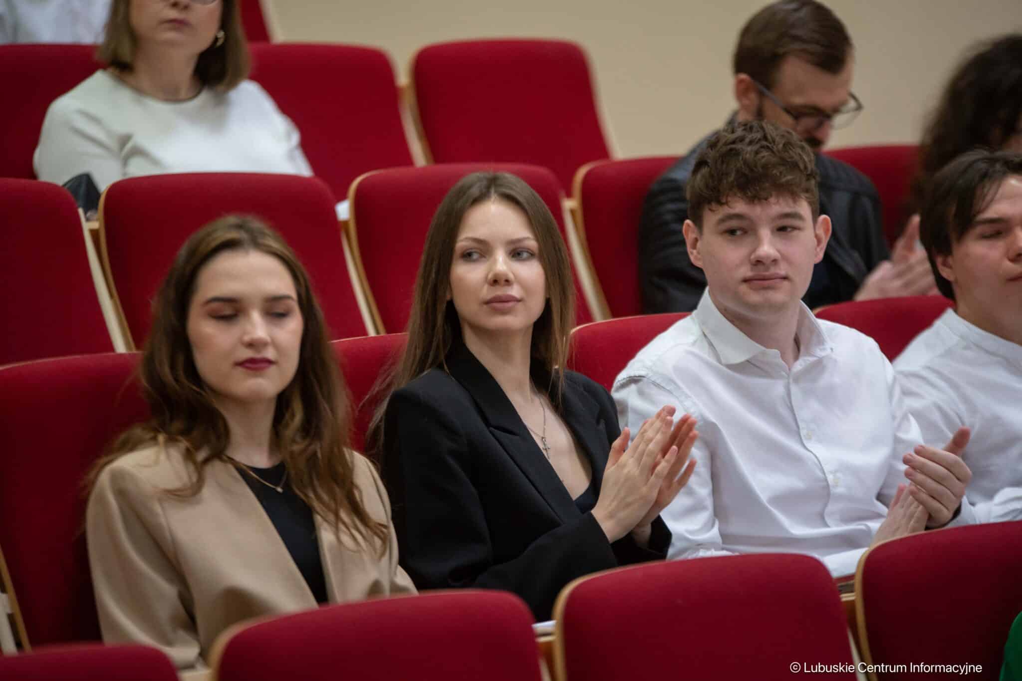 Three young adults seated in red theater seats, with two women in blazers at center-left and a man in a white shirt on the right, all clapping as an audience.
