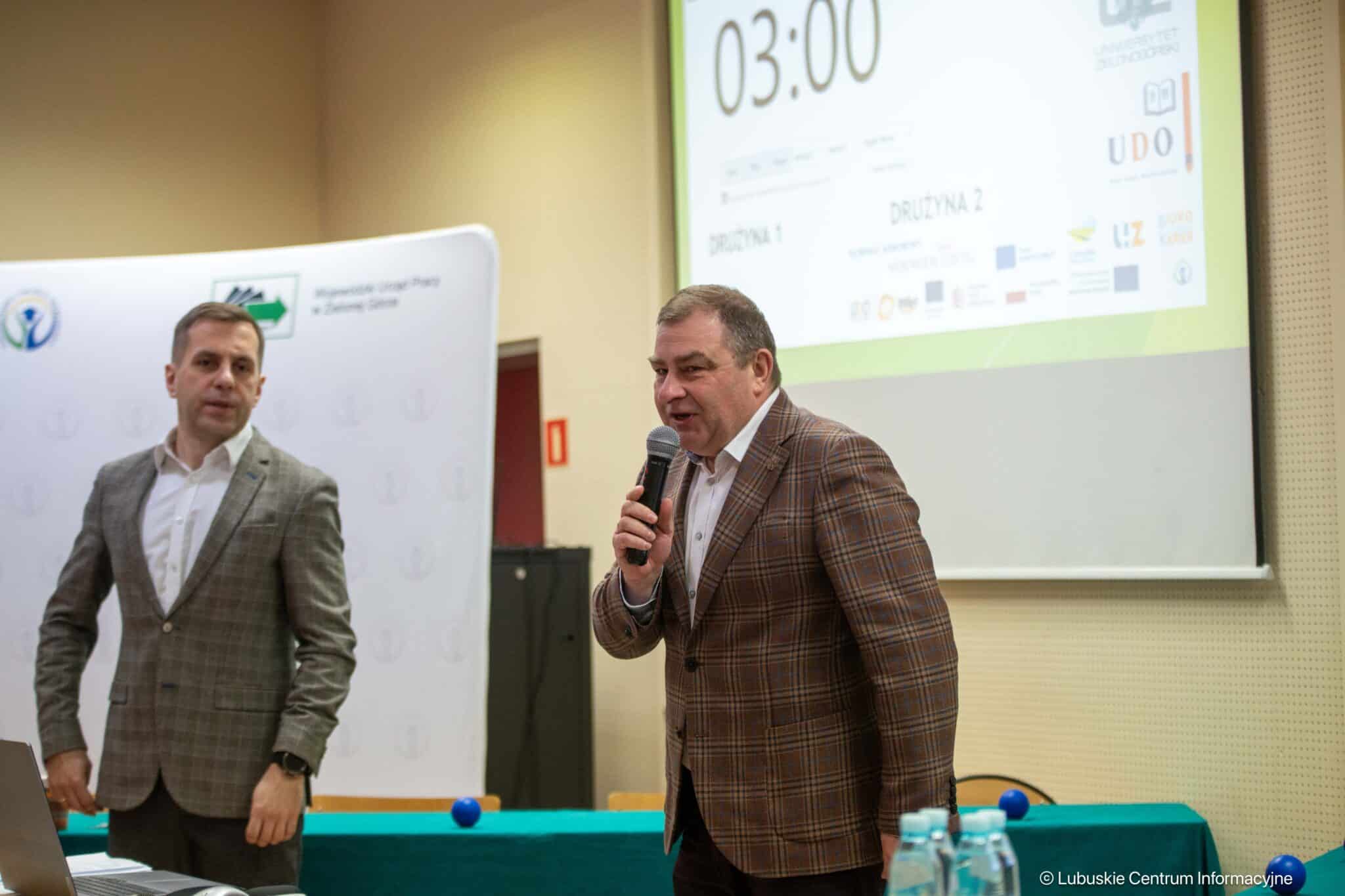 Man in a brown plaid blazer speaks into a handheld microphone at a conference, while another man in a gray suit stands beside a laptop on a table in a meeting room.