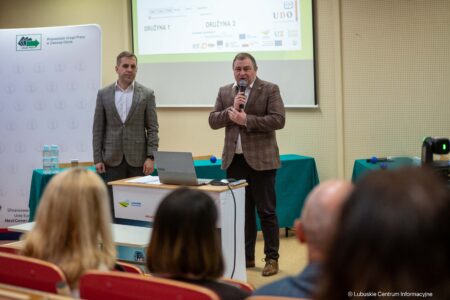 Two men in business suits present at a conference; one speaks into a microphone near a laptop on a podium.