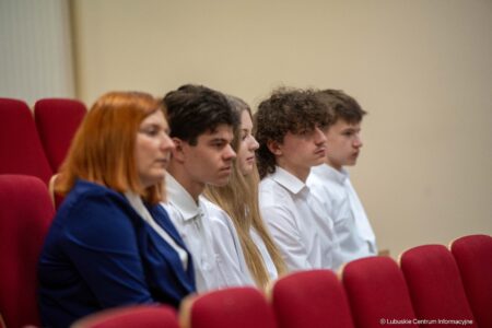 Group of five students seated in red auditorium chairs, listening attentively; woman with red hair in a blue jacket in the foreground and others in white shirts behind her.