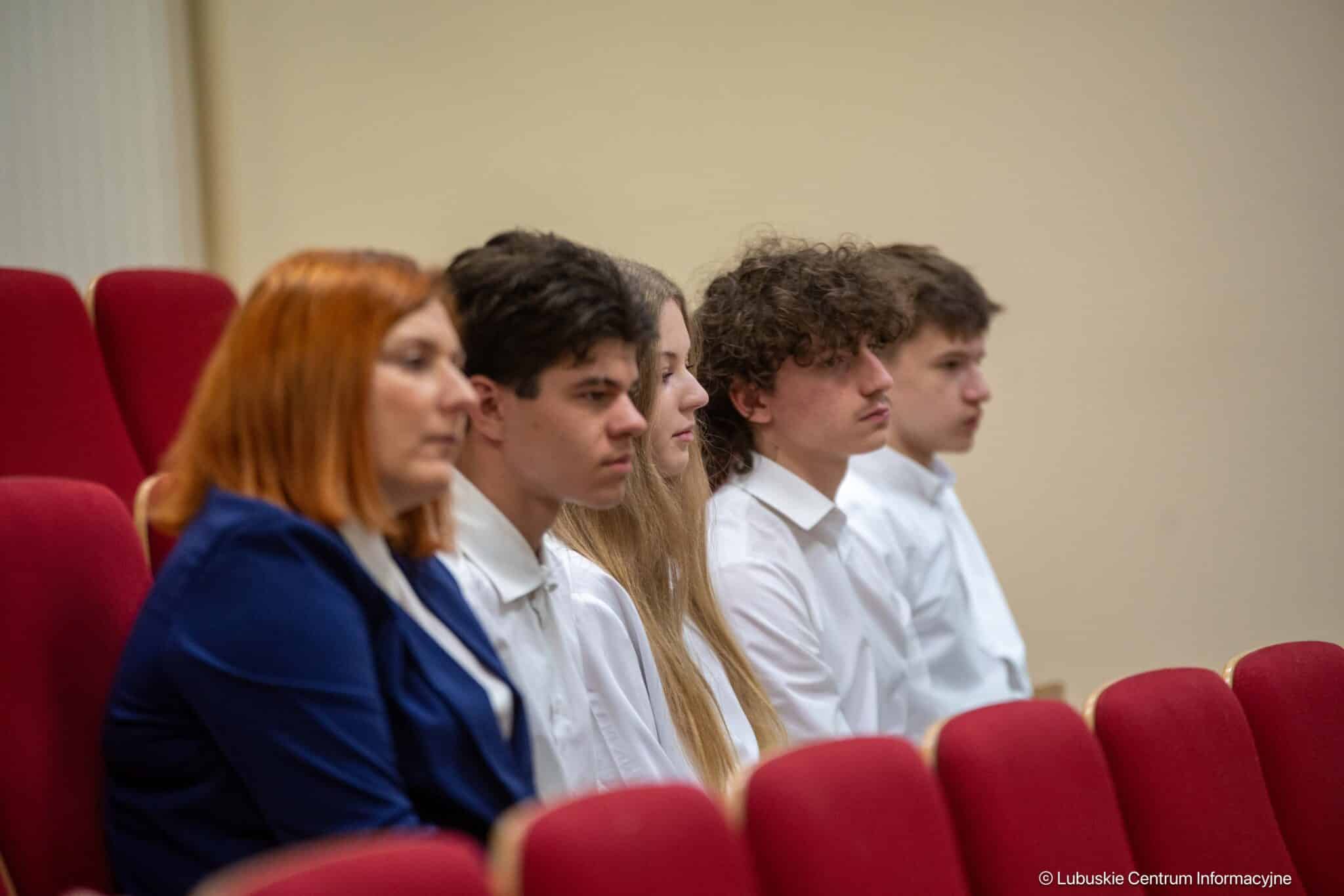 Group of five students seated in red auditorium chairs, listening attentively; woman with red hair in a blue jacket in the foreground and others in white shirts behind her.