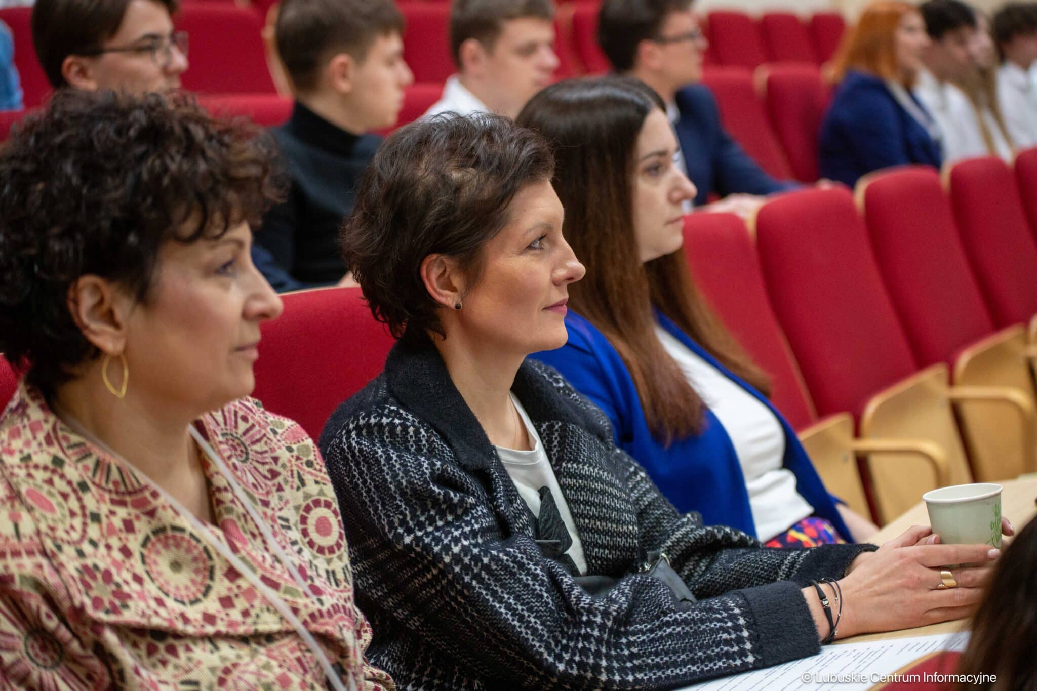 Audience seated in red theater chairs listening to a presentation in a conference or lecture hall.