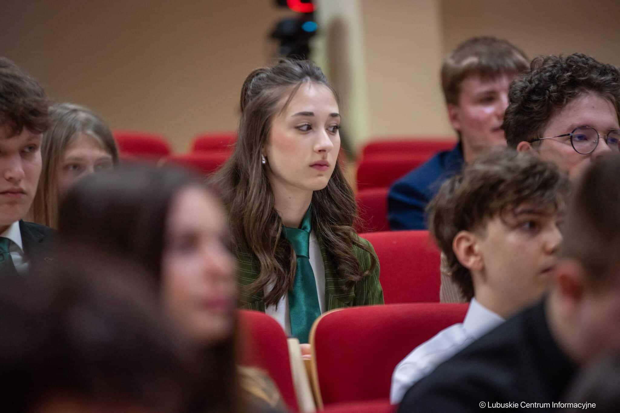 Young woman in green blazer and teal tie sitting in an auditorium among peers, looking ahead thoughtfully.