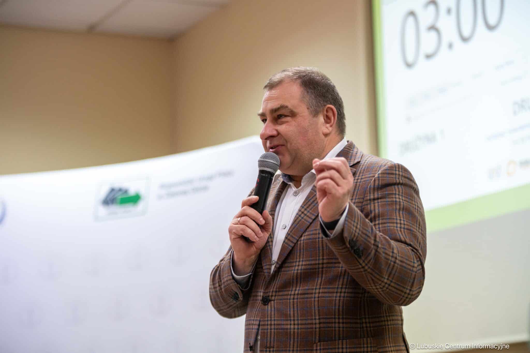 Man in a brown plaid blazer speaks into a handheld microphone during a conference or presentation on stage.