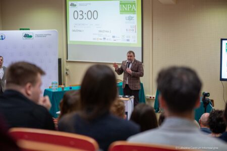 Speaker in a brown plaid blazer addressing an audience from a podium in a conference room, with a large timer screen behind him (03:00) and banners along the wall.