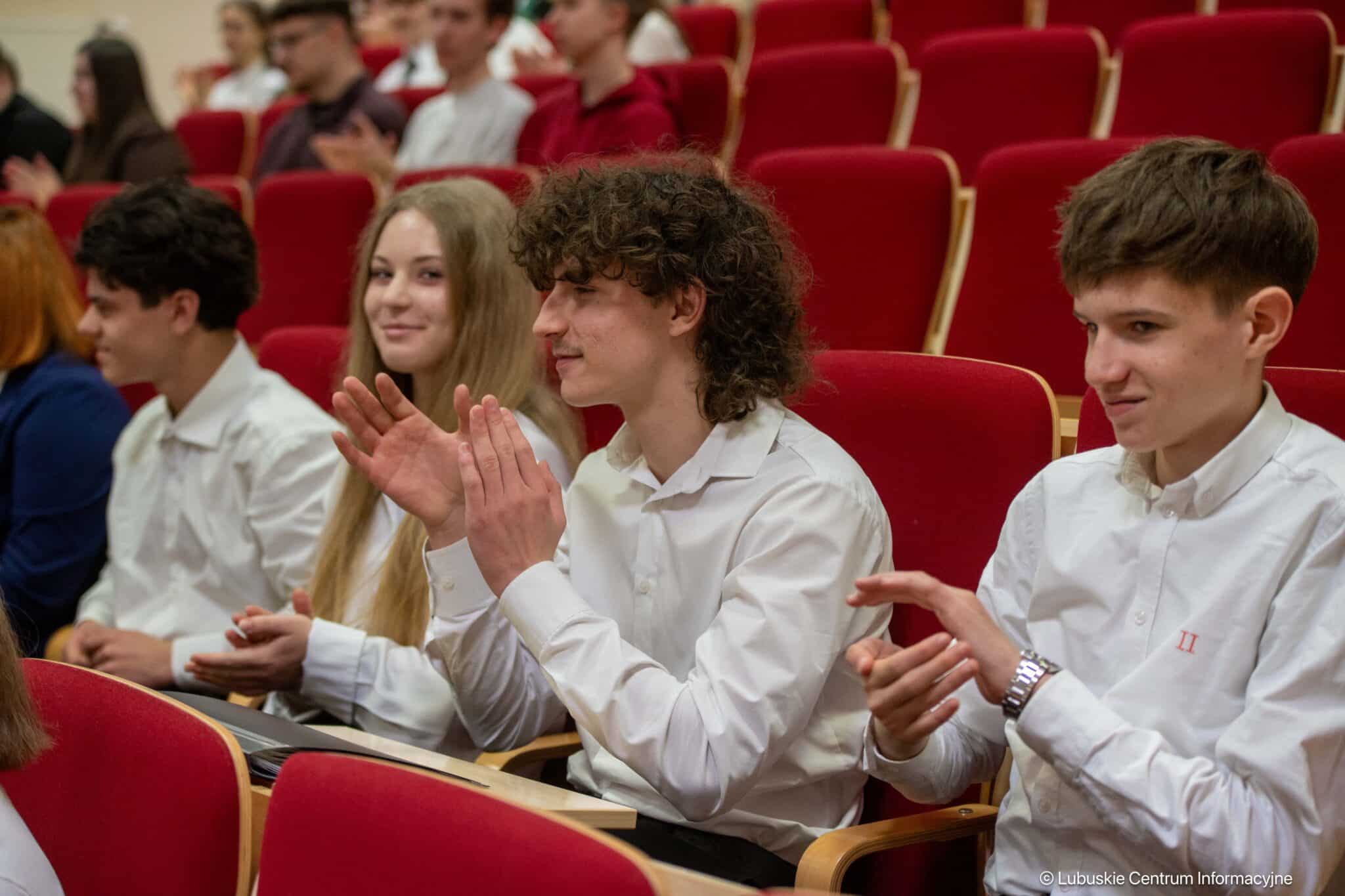 Students in white shirts sit in red theater seats clapping during a ceremony or presentation  Słowna szermierka w oksfordzkim stylu