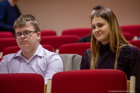 Two young adults sit in a red theater, the man with glasses on the left has eyes closed and a white shirt, the woman on the right smiles slightly.