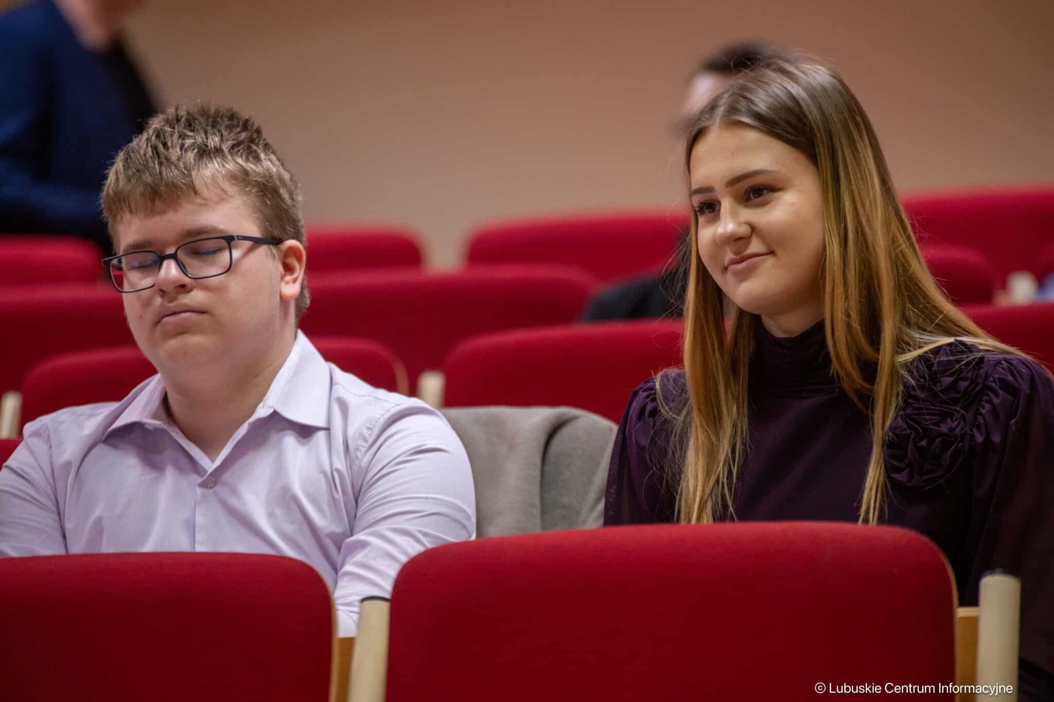 Two young adults sit in a red theater, the man with glasses on the left has eyes closed and a white shirt, the woman on the right smiles slightly.