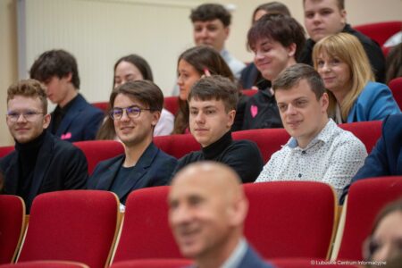 Group of young adults seated in a red-velvet auditorium, some smiling and chatting while paying attention to an event.