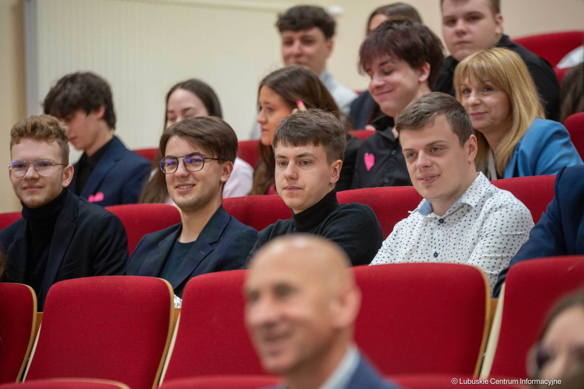 Group of young adults seated in a red-velvet auditorium, some smiling and chatting while paying attention to an event.