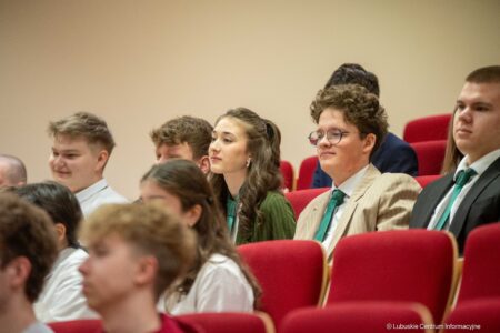 Group of young students seated in red auditorium chairs, paying attention at a formal event in a classroom or lecture hall.