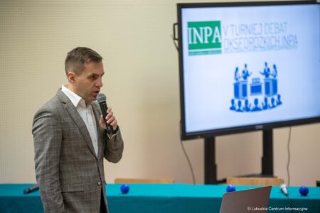 Man in a gray checkered blazer speaks into a handheld microphone during a presentation beside a screen displaying the INPA logo.