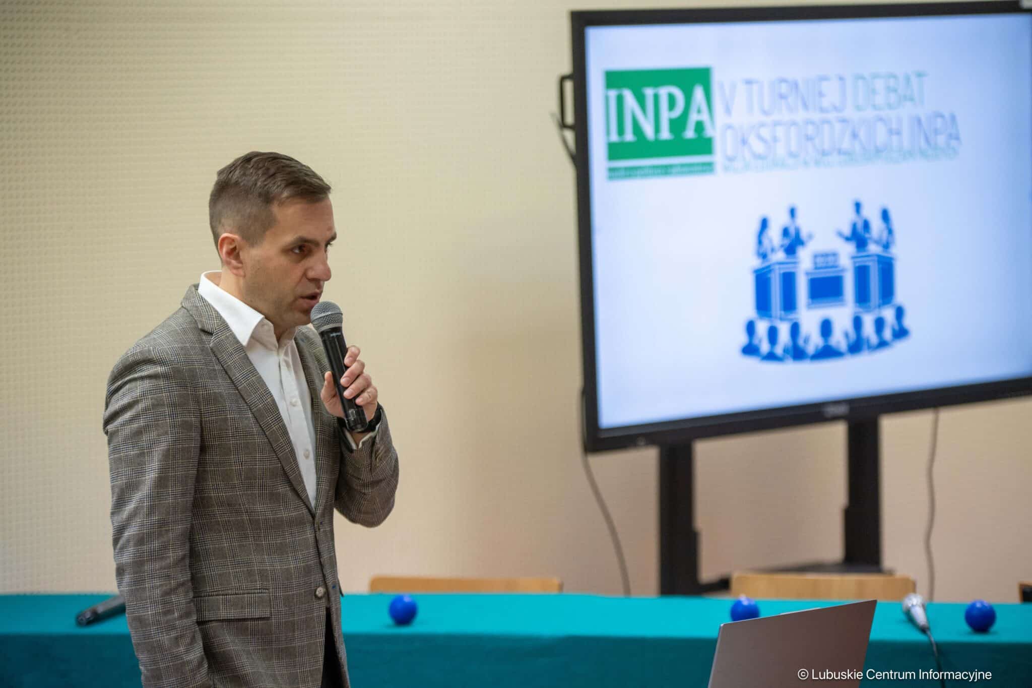 Man in a gray checkered blazer speaks into a handheld microphone during a presentation beside a screen displaying the INPA logo.