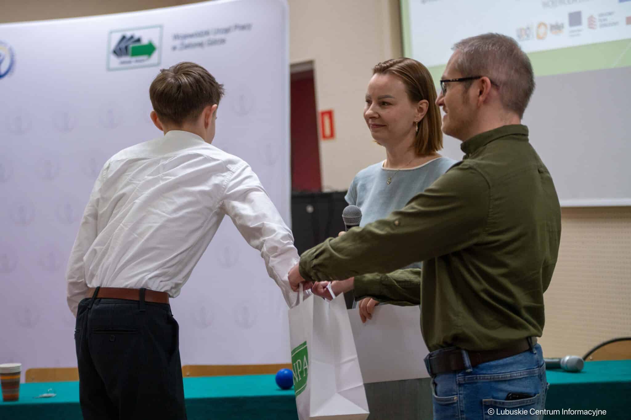 People at an event: a man in a white shirt shakes hands and receives a gift bag from a man in a green shirt, while a woman with a microphone watches.
